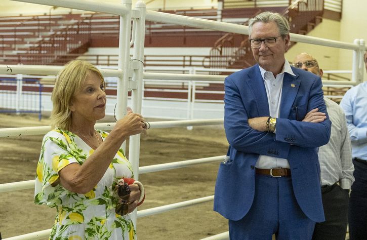 A woman in a floral dress gestures while talking to a man in a blue suit with folded arms. They are standing near white metal railings in what appears to be an indoor arena with empty bleachers.