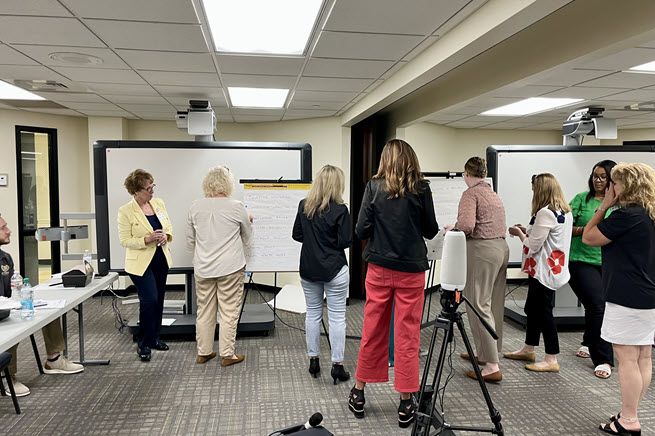 Eight women face away from the camera towards flip chart easels at the front of a conference room. They have sticky dots in their hands and are voting on what's most important to them.
