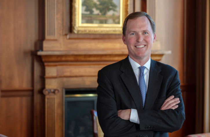 A man in a checked dark suit and light blue shirt stands indoors, smiling, with his hands resting on a wooden railing in a modern office lobby with soft lighting.