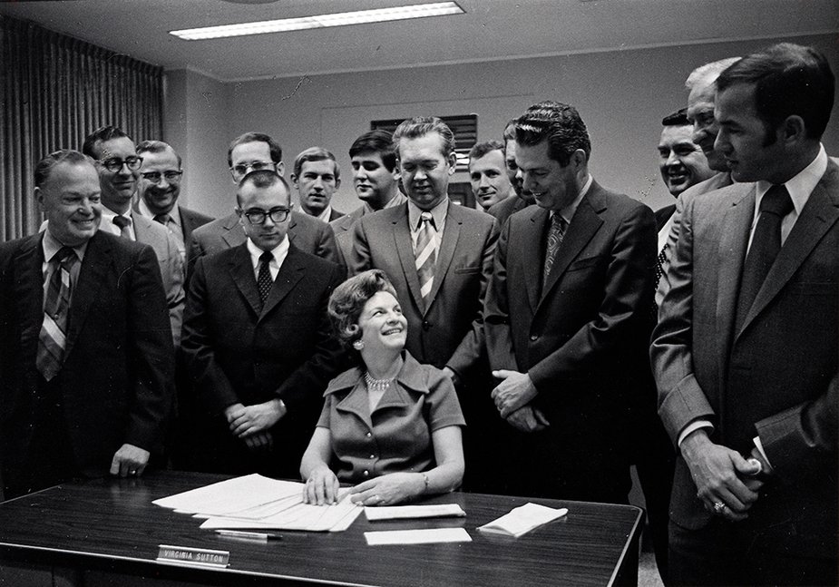 Image Description. Black and white photograph shows a Caucasian woman siting at a table while a dozen men stand around her.