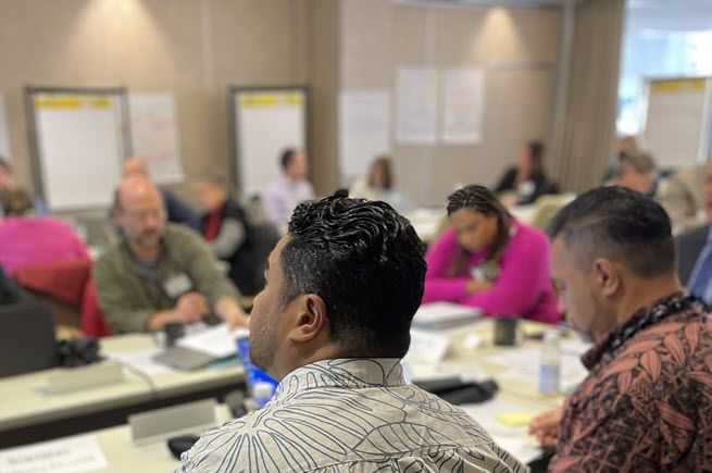 The photo is of a roomful of people working in small groups. The two men closest to the photographer are in high definition, and everyone beyond them is blurred.