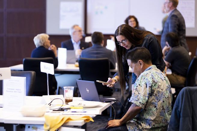 A white woman with long brown hair is holding a cell phone. She leans toward a dark-haired man from Hawaii wearing a floral shirt and sitting in front of his laptop. They appear to be checking something online.