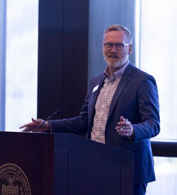 Tall white man with a beard and reddish-gray hair stands at the podium. He's in a blue suit and striped shirt. He looks comfortable and relaxed, like someone just cracked a joke.
