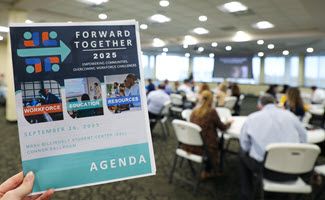 A woman's hand is holding the Forward Together 2025 agenda, in teal, blue and red. The agenda is in high definition, while the room full of participants working at round tables are blurry and distant.