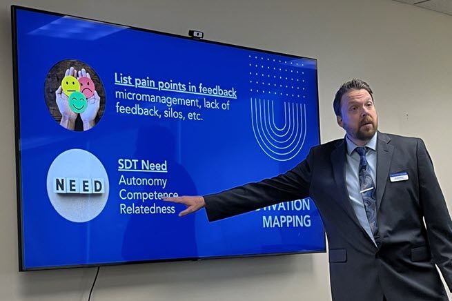 A White man in a blue suite with dark hair and a beard stands in front of a digital display mounted on the wall, and points to words like autonomy and relatedness that are part of emotional intelligence.