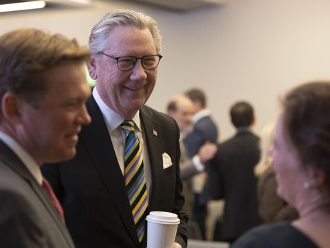 Three people in business attire are conversing in a conference or meeting room. The man in the center is smiling and holding a coffee cup, while others engage in conversation in the blurred background.
