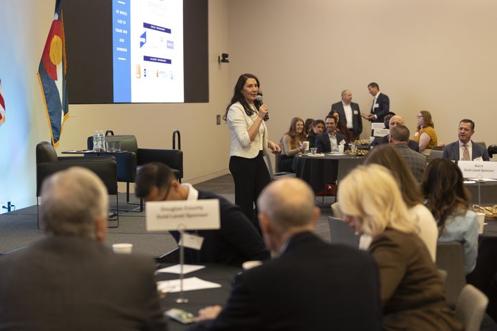 A woman speaks into a microphone on stage at a conference, with people seated at round tables listening. A presentation slide is projected behind her and a state flag stands to her left.