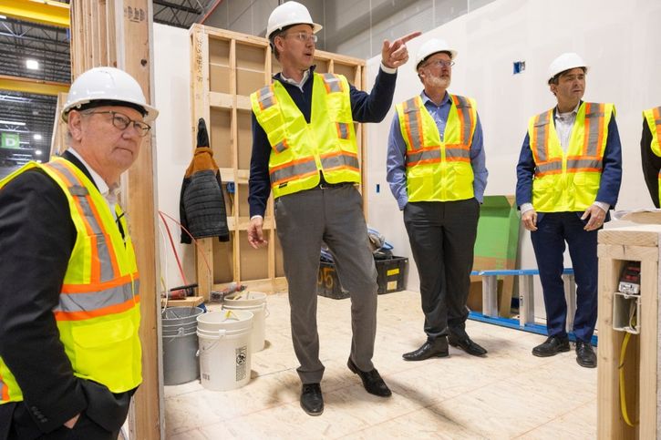 Four men wearing hard hats and yellow safety vests stand indoors at a construction site. One man gestures while speaking, and building materials and tools are visible in the background.