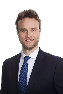 Head shot of a white man around 40 in a dark suit with a blue tie.