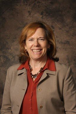 A woman in her 50s with red hair, a maroon short and gray jacket stands with her arms crossed and a smile on her face.
