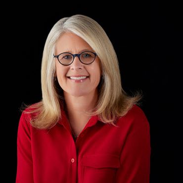 A White woman in her 40s is smiling at the camera. She has shoulder-length blonde hair and is wearing oval glasses and a bright red shirt, which looks dramatic against a black background.