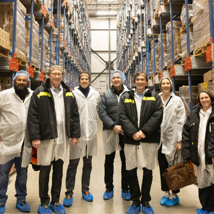 A group of people wearing protective coats, blue shoe covers, and some with hairnets stand smiling in a warehouse aisle lined with tall shelves stacked with boxes.
