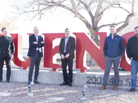 Five men stand in front of large red letters spelling “BUENO” outdoors, with leafless trees and a building in the background. The group is smiling and dressed in business-casual attire.