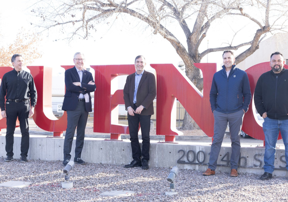 Five men stand in front of large red letters spelling “BUENO” outdoors, with leafless trees and a building in the background. The group is smiling and dressed in business-casual attire.
