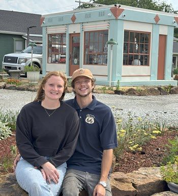 A couple in their 20s sit on a rock wall in front of a 1920s gas station. She has long, strawberry blonde hair and he's wearing a ball cap and a Route 66 t-shirt.