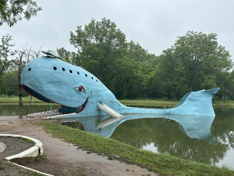 A large blue whale sculpture sits partially in a pond, with its mouth open and steps leading inside. Trees and greenery surround the whimsical, cartoon-like structure on a cloudy day.