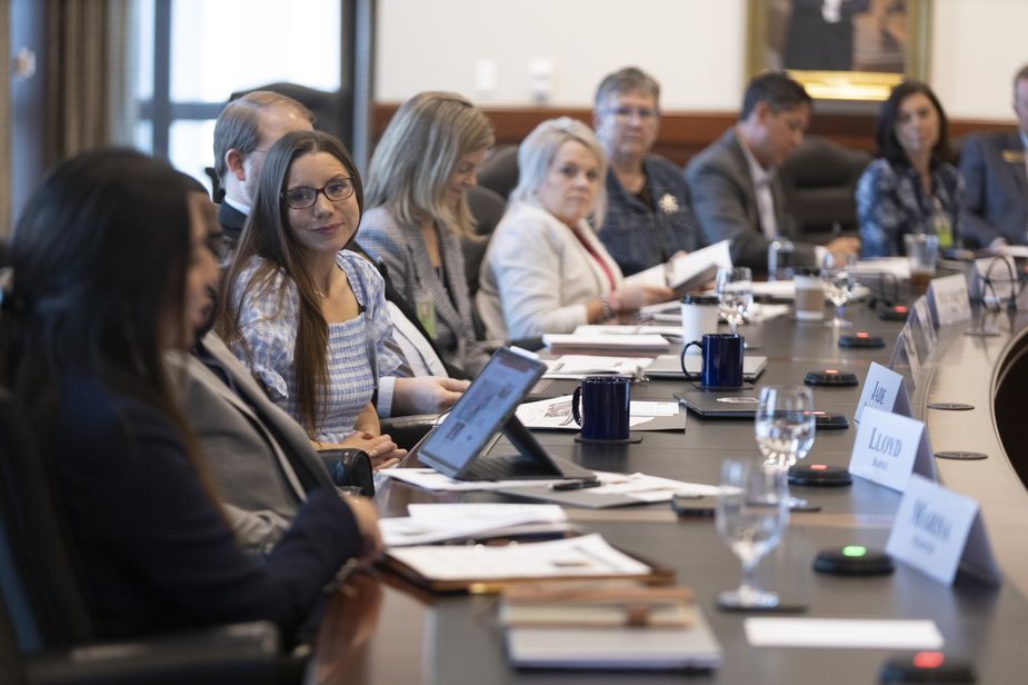 A young woman turns towards the camera, in conversation with her neighbor. She is along a row of about eight people along the side of a big wooden conference table.