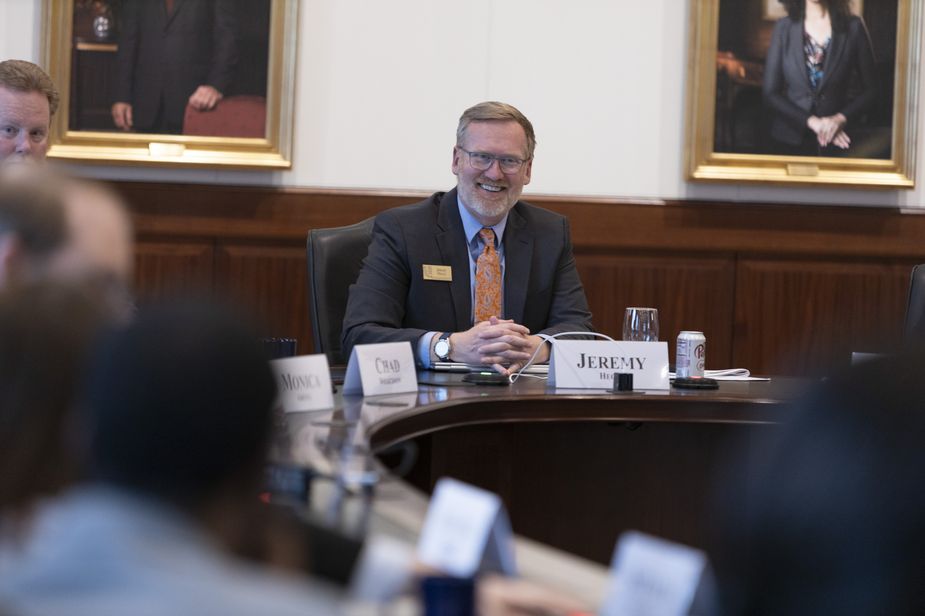 At the head of the conference table, a smiling, bearded White man in a dark suit and red tie, faces the camera.
