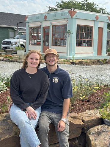 A young couple sit on a rock wall about 100 yards in front of an old service station that they have restored.