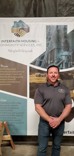 A smiling White man in a gray polo shirt is standing in front of a vertical banner celebrating Interfaith Housing, a nonprofit in Hutchinson, Kansas.