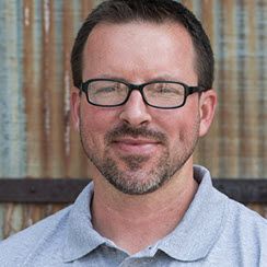 Photo shows a White man in his 40s with a short beard and mustache, wearing oblong-framed glasses. He has dark brown hair and is wearing a light blue shirt.