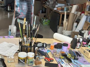 Paintbrushes, paints, scissors and other artist supplies cluster on a table.