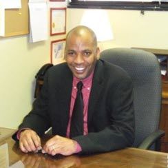 The photo shows a 40-something African American man in a dark suit and red shirt sitting at his desk. He is smiling.