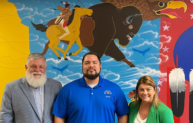 Two men and a woman pose in front of a Native American-inspired mural with a buffalo being chased by a young man on a horse.