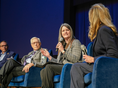 Four people sit in blue chairs on stage during a panel discussion. One woman holds a microphone and speaks while the others listen attentively. The background is blue and they appear to be at a formal event.