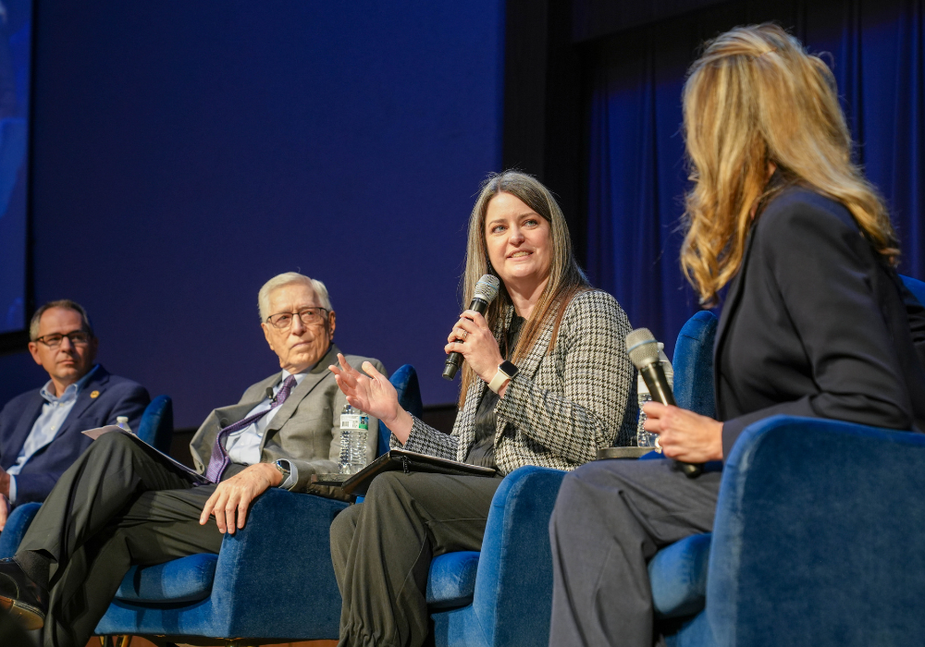 Four people sit in blue chairs on stage during a panel discussion. One woman holds a microphone and speaks while the others listen attentively. The background is blue and they appear to be at a formal event.