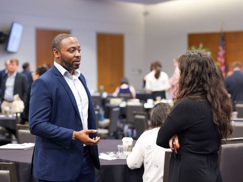 A Black man in a navy blue suit and white shirt talks to a woman with long brown hair, wearing a black sweater. It looks like the conference is on break.