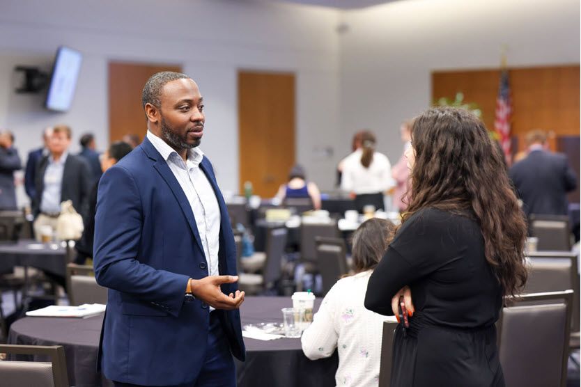 A Black man in a navy blue suit and white shirt talks to a woman with long brown hair, wearing a black sweater. It looks like the conference is on break.
