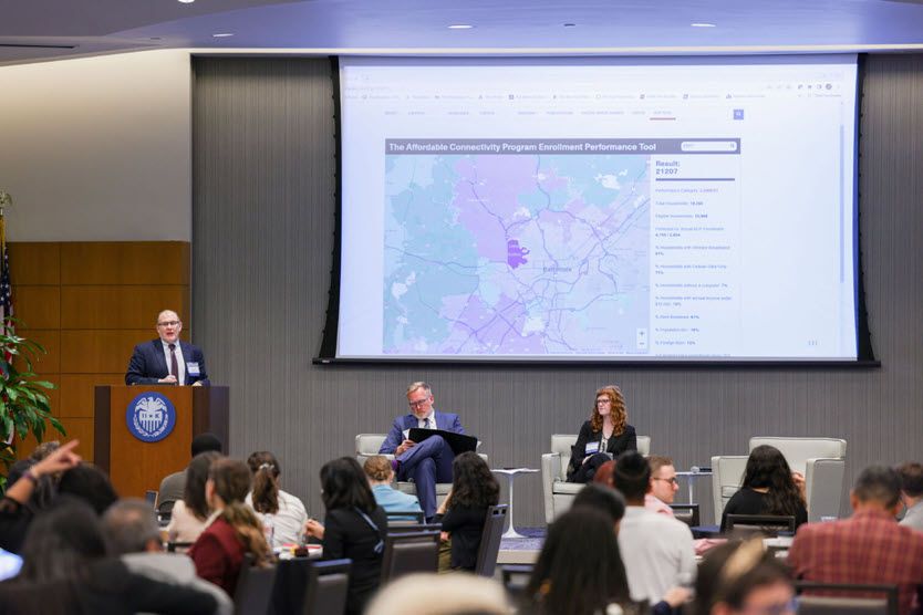 Three white people on the stage. The older man stands at the podium, and a man in his 40s and a woman in her 30s occupy chairs on the stage.