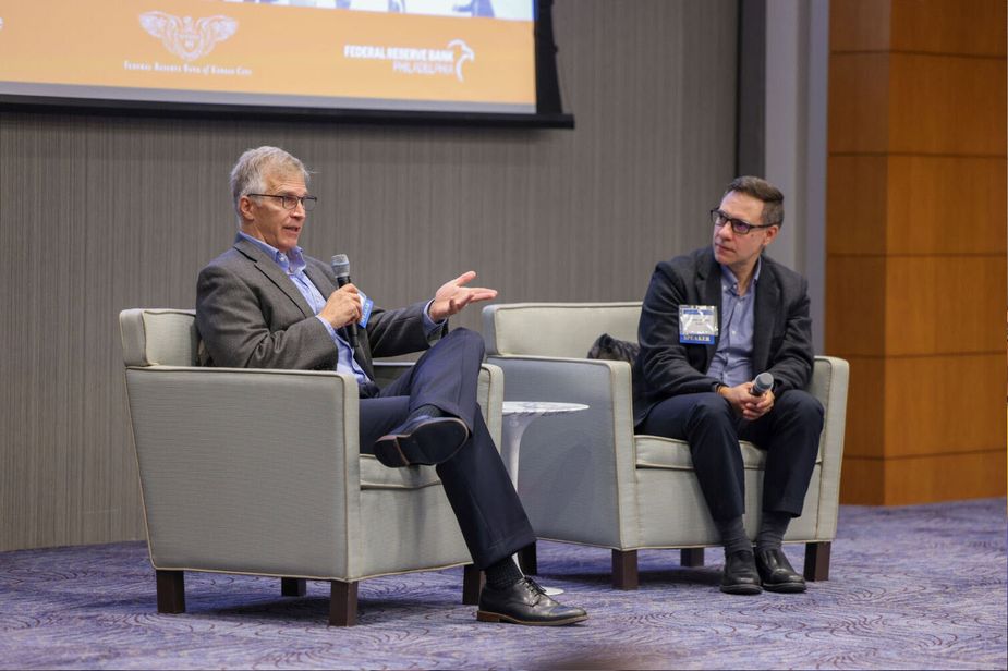Two men sit on overstuffed chairs on a stage. Both are white. The one with the blonde hair is speaking, the one with brown hair is listening.