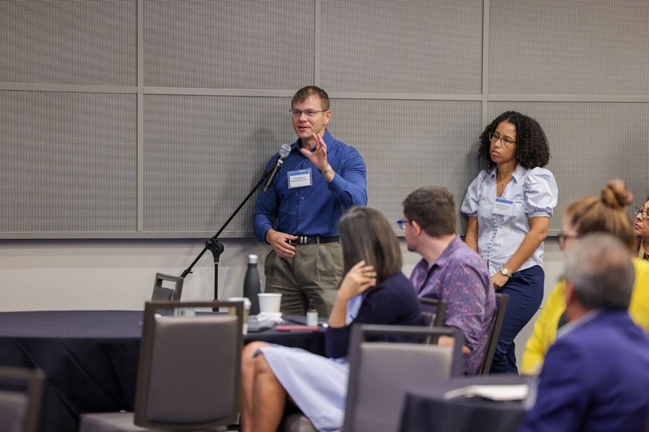 You can't see them, but there are presenters on the stage. Out in the audience, two people stand near a microphone that has been set up for people who have questions. The man talking into the microphone is in his 40s, white, and wearing a bright blue shirt and khaki pants.