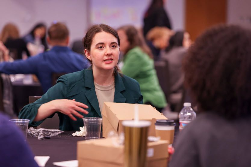 A White woman in her 20s opens her boxed lunch as she talks with the Black woman across the table from her.