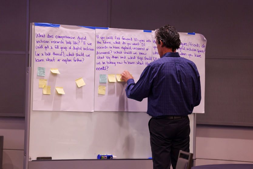 A white man with short brown hair, wearing a blue cotton shirt, stands with his back to the audience. He is adding Post-It notes to a flip chart page that contains writing.