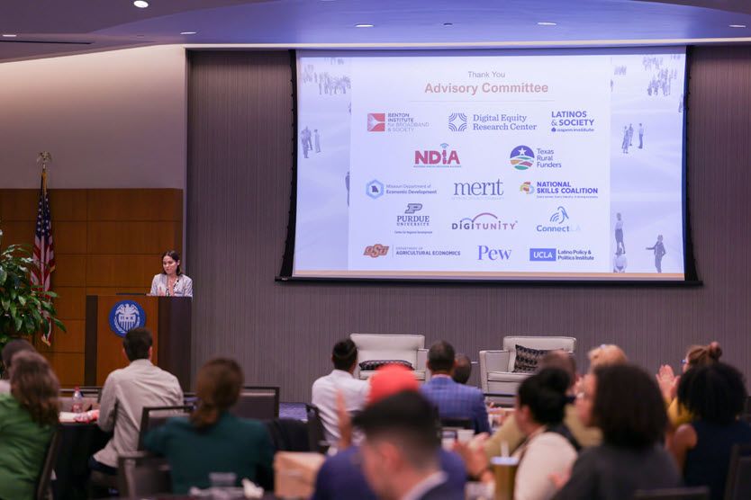 A white woman in her 30s stands at a podium, facing the audience. Behind her is a slide showing all the members of the event planning committee.