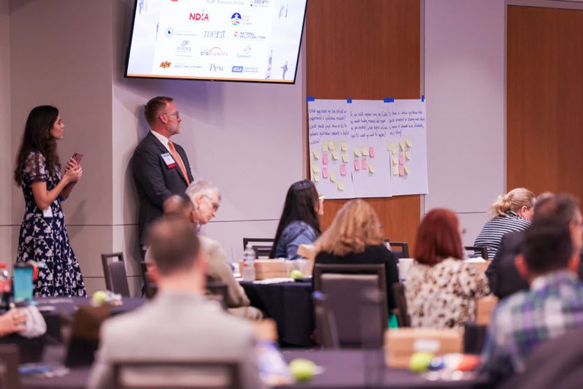 We can't see the person, but someone is speaking from the stage. We see two people, a woman and a man, standing against the wall, listening. Other people are seated at tables facing the stage.