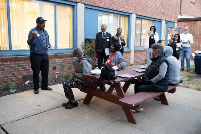 A Black man in a brown jacket and baseball cap smiles as he talks to people sitting on a picnic bench and standing nearby.