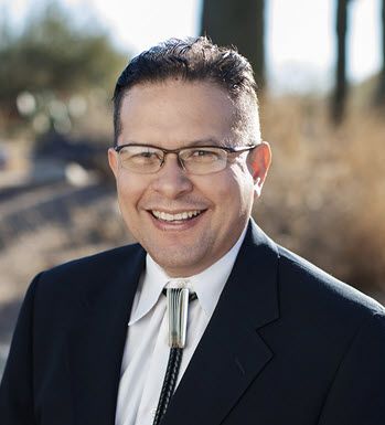 Photo is of a dark-haired man in a black suit and silver tie.