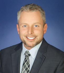 The photo is a headshot of a 30-something White man wearing a gray suit and a blue shirt, and smiling at the camera.
