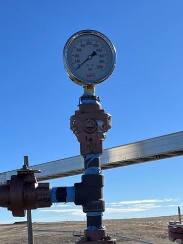 A round, chrome gauge on an orange metal stand is highlighted against a bright blue sky and brown Kansas prairie.