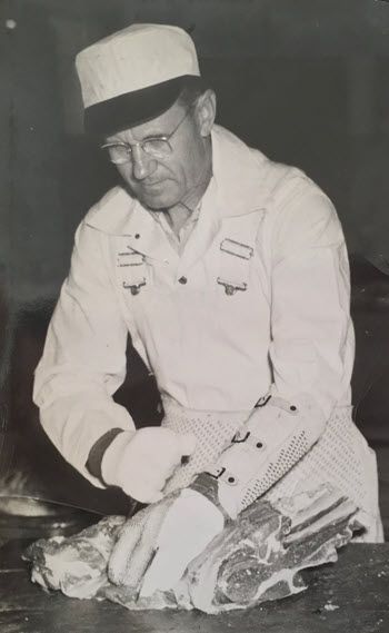 A trim, powerful-looking white man in his 50s wears a white cap and jumpsuit and thick glasses. He stands over a large cut of beef on a counter, and uses twine to hold it together.