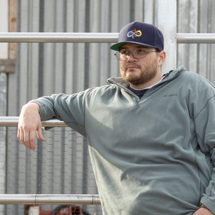 A man in a gray sweatshirt and dark cap leans against a metal fence, with a corrugated metal building and a window in the background. He has glasses and a beard, looking off to the side with a relaxed posture.