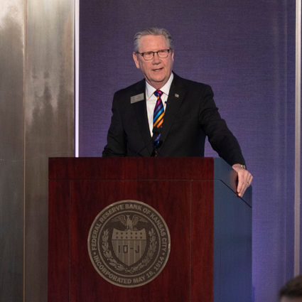 A man in a suit and striped tie speaks at a wooden podium with an official seal, in front of blue and gray curtains at an indoor event.