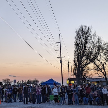 A crowd gathers at dusk near a glowing neon Hi-Way Cafe sign, with trees, power lines, and small buildings visible in the background.