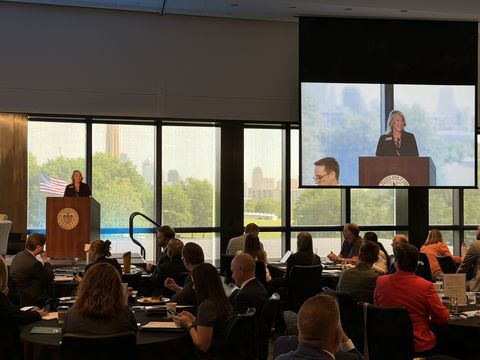 A woman speaks at a podium in front of a seated audience in a conference room, with large windows showing greenery outside. Her image is displayed on a large screen beside her.