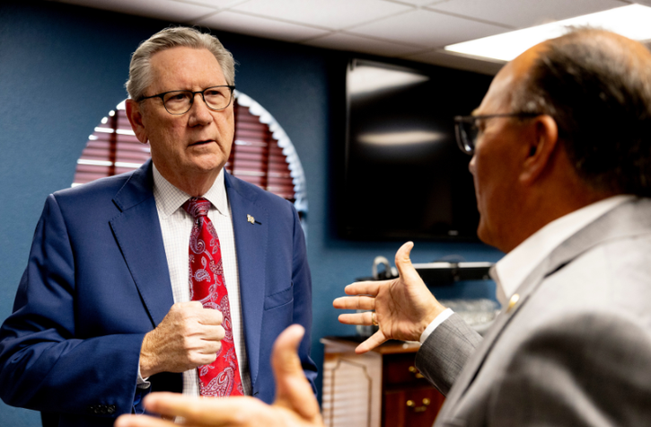 Two men in suits have a serious conversation in an office. One man gestures with his hands while the other listens intently, appearing thoughtful, with a fist near his chest. A TV and wood blinds are visible in the background.
