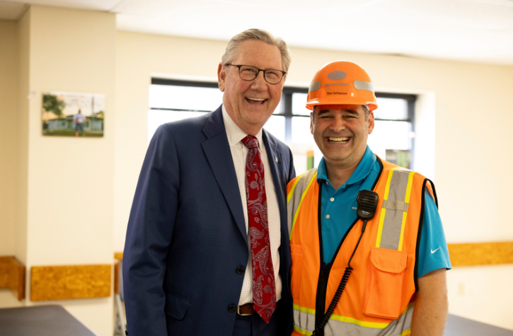 Two men smiling indoors; one wears a blue suit and red tie, the other wears a construction vest, hard hat, and has a radio clipped to his vest.
